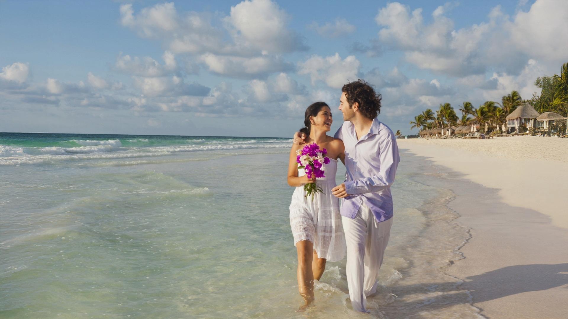 Smiling couple walks along sandy beach, woman holding purple flowers