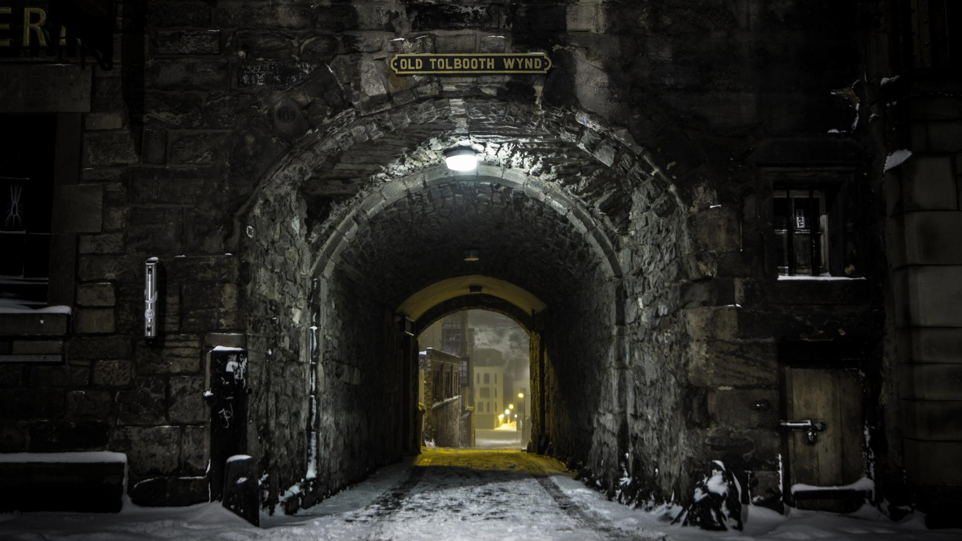 Stone archway at night leading to a narrow street with lit buildings in the distance.