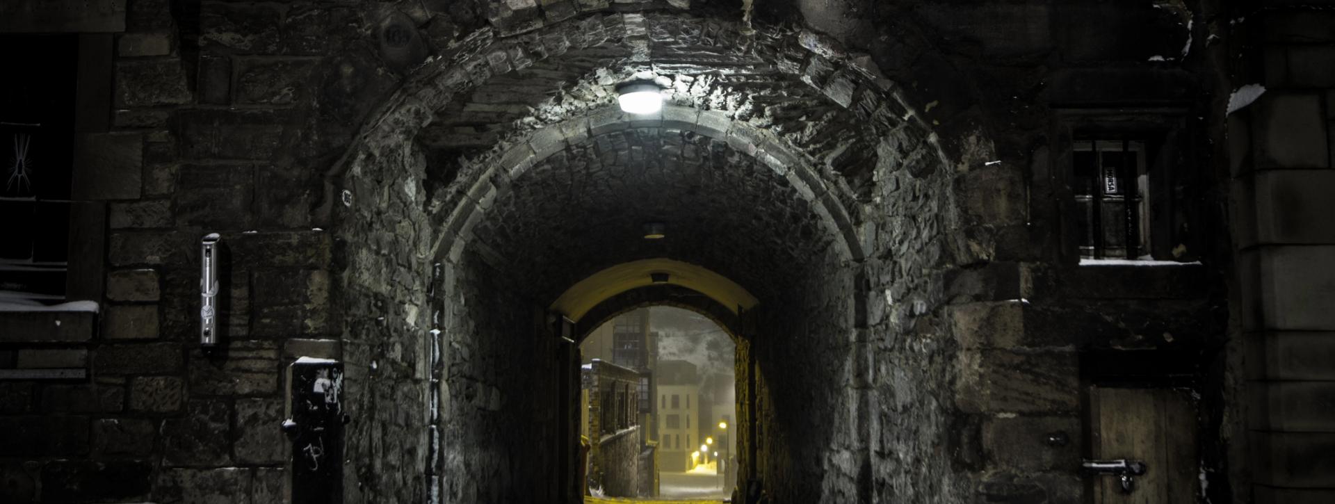 Stone archway at night leading to a narrow street with lit buildings in the distance.