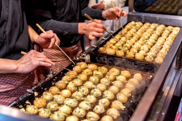 Hands turning rows of takoyaki balls on a hot grill at a food stall