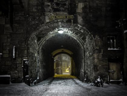 Stone archway at night leading to a narrow street with lit buildings in the distance.