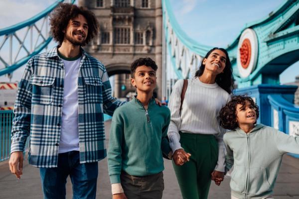 A smiling family of four (two parents, two children) walking across a bridge, with the Tower Bridge in London visible in the background.