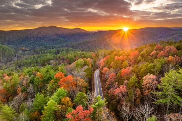 Sunset shining over forested mountains with a winding road through autumn trees.