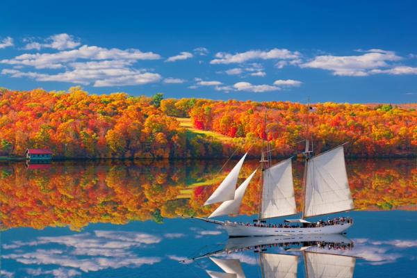 White sailboat gliding across a calm lake with colorful autumn trees in the background.