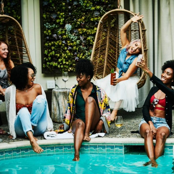 Group of women enjoying drinks and taking selfies while sitting by a stylish resort pool during a fun girls' trip.