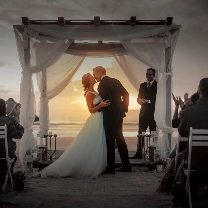Bride and groom kiss under wedding canopy on beach at sunset with guests