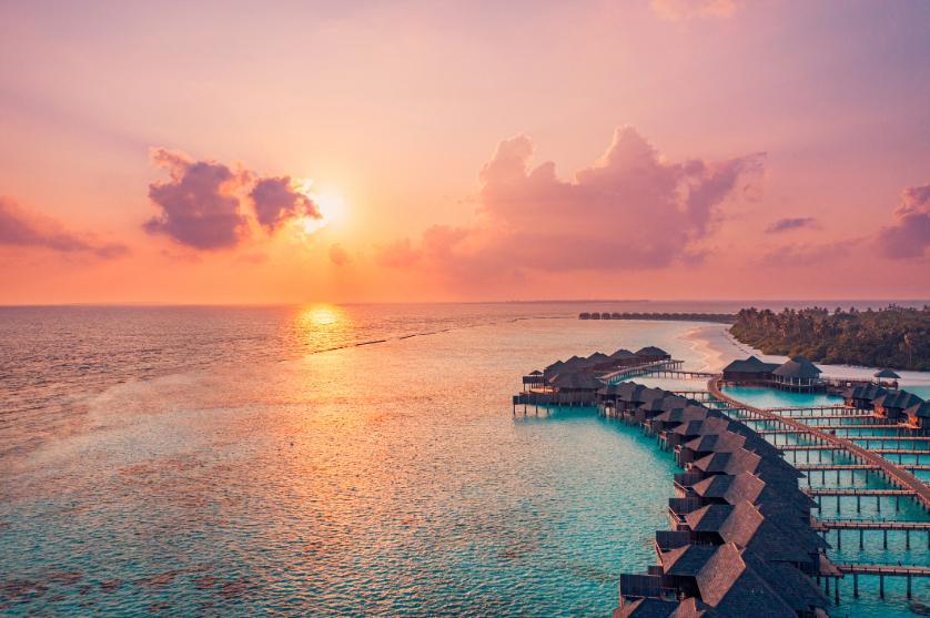 High-angle view of thatched-roof overwater villas on a curving pier during a vibrant orange and pink sunset over the calm ocean.