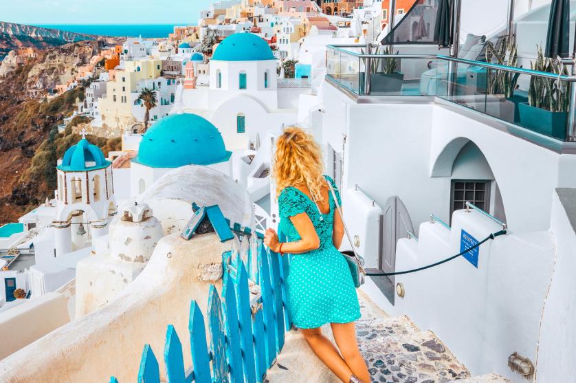 A woman in a teal dress looking out over the white and blue cliffside buildings of Santorini, Greece, with the Aegean Sea in the distance.