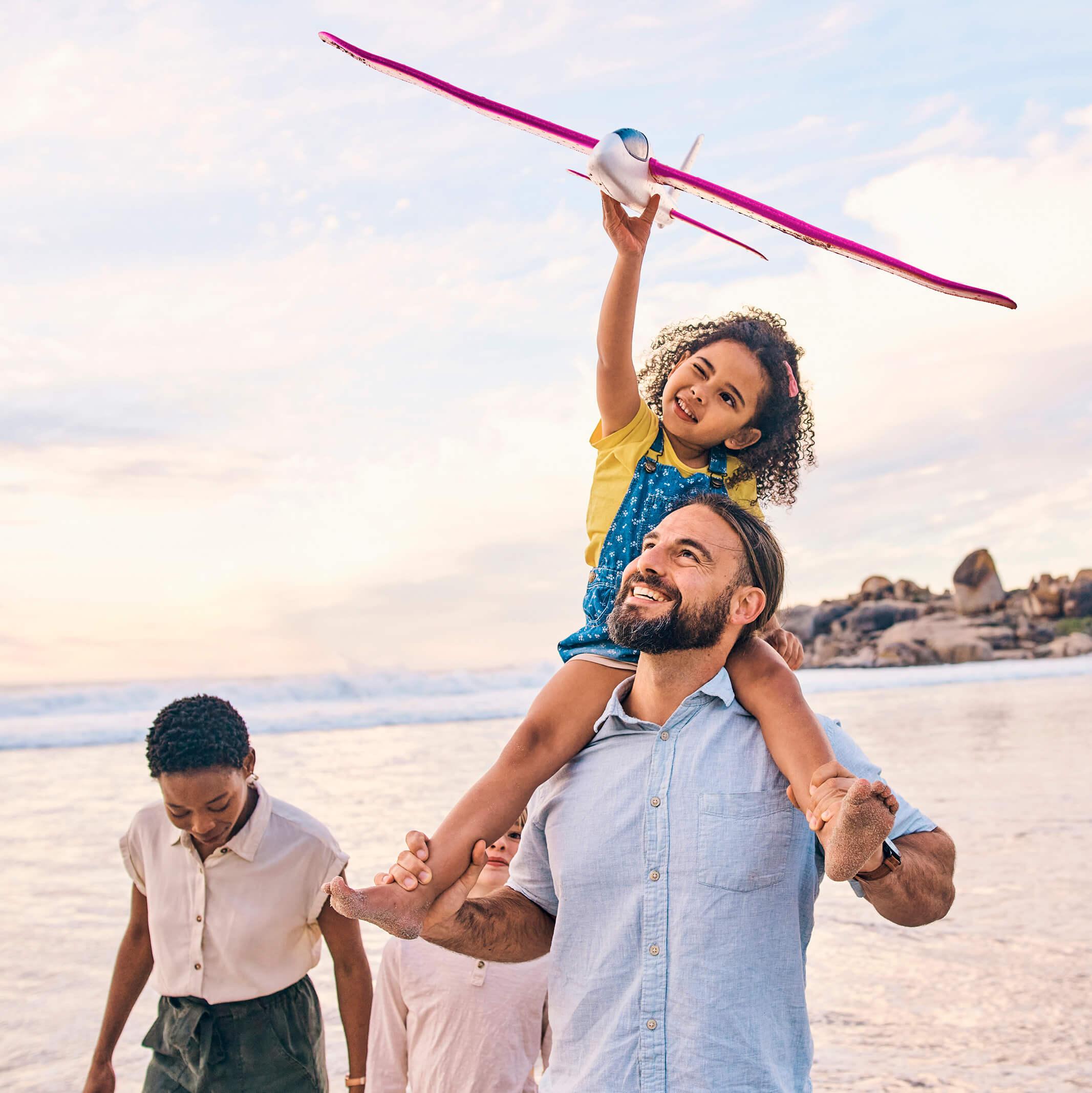 A family walks along the shore, a daughter on her father's shoulders while flying a toy airplane