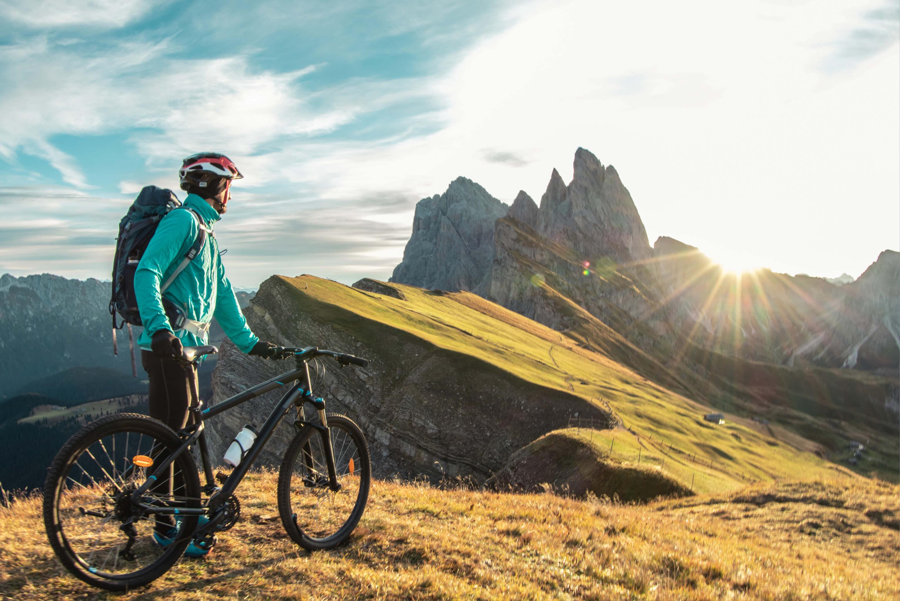 A man stands with a bike on a hill, gazing at the mountains in the distance, where the sun sets