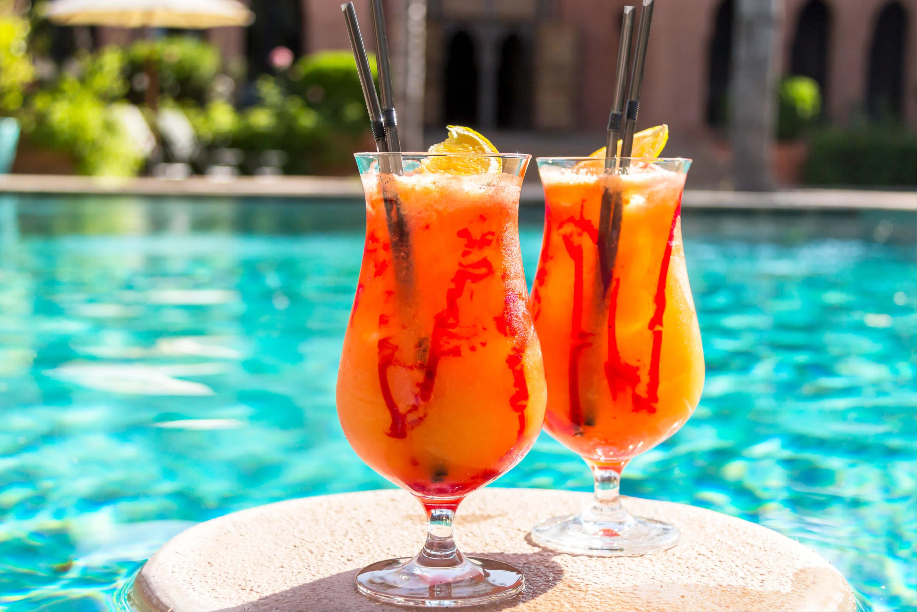 Two refreshing orange tropical drinks in front of a shimmering swimming pool