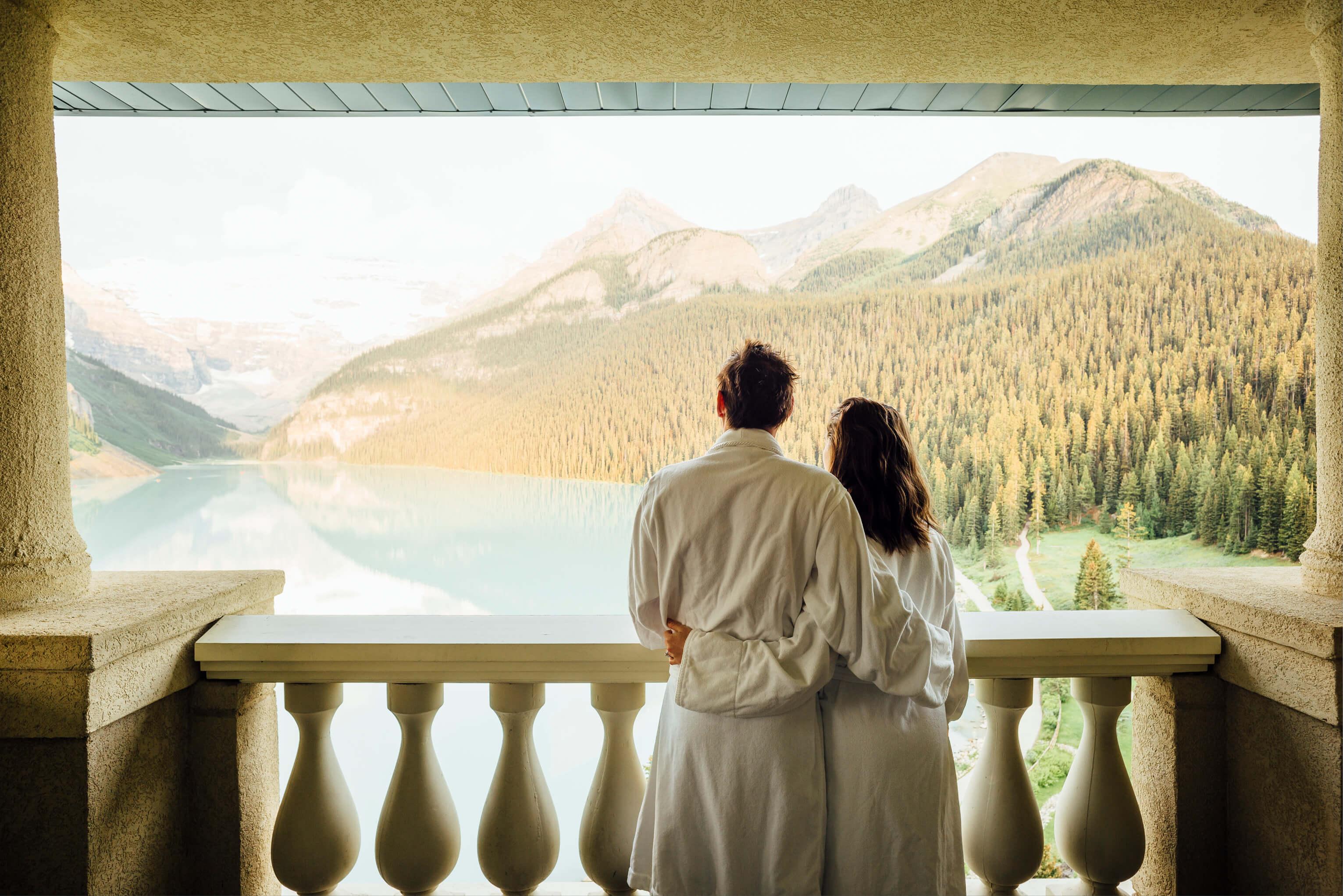 A couple in robes gazes at a serene lake surrounded by mountains, enjoying a peaceful moment together