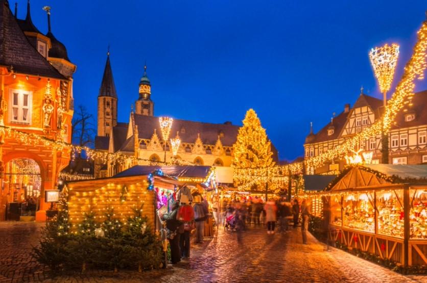 Christmas market in Europe illuminated with festive lights, decorated stalls and a towering Christmas tree at dusk.
