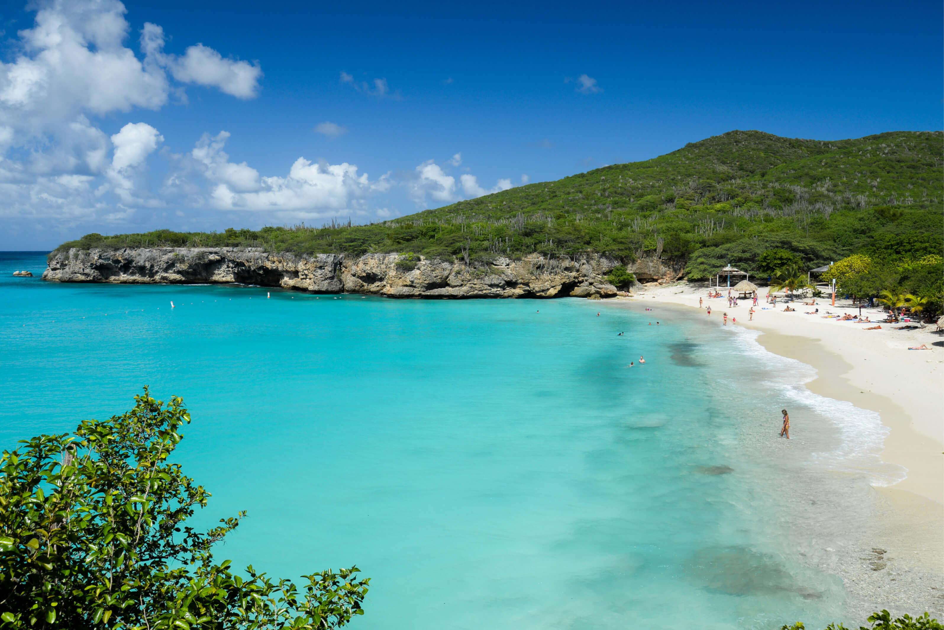 A sandy beach in Curacao stretches along a clear blue ocean under a bright sky, inviting sunbathers and swimmers