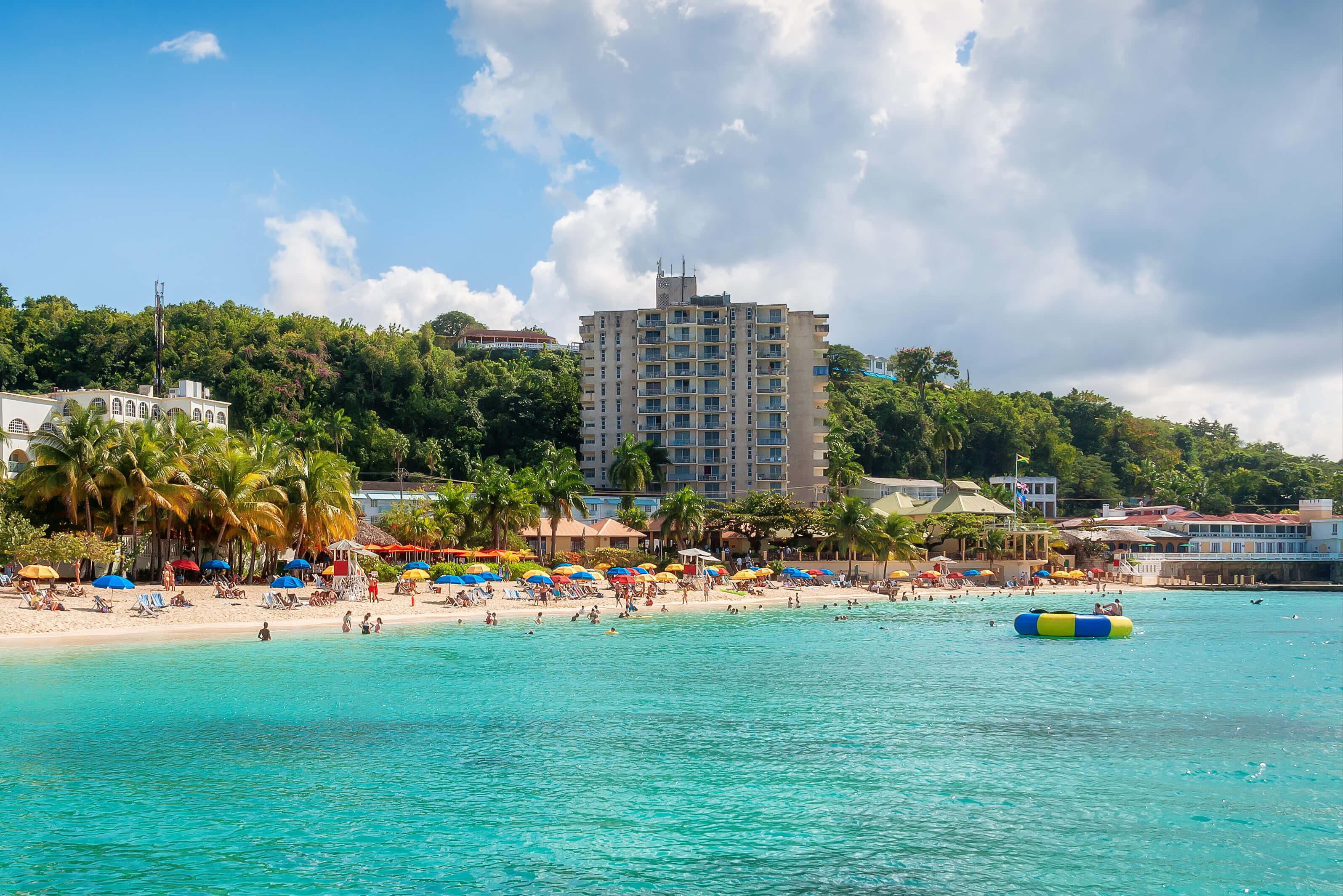 A lively beach scene in Montego Bay, Jamaica with numerous people enjoying the sun, alongside a hotel in the background