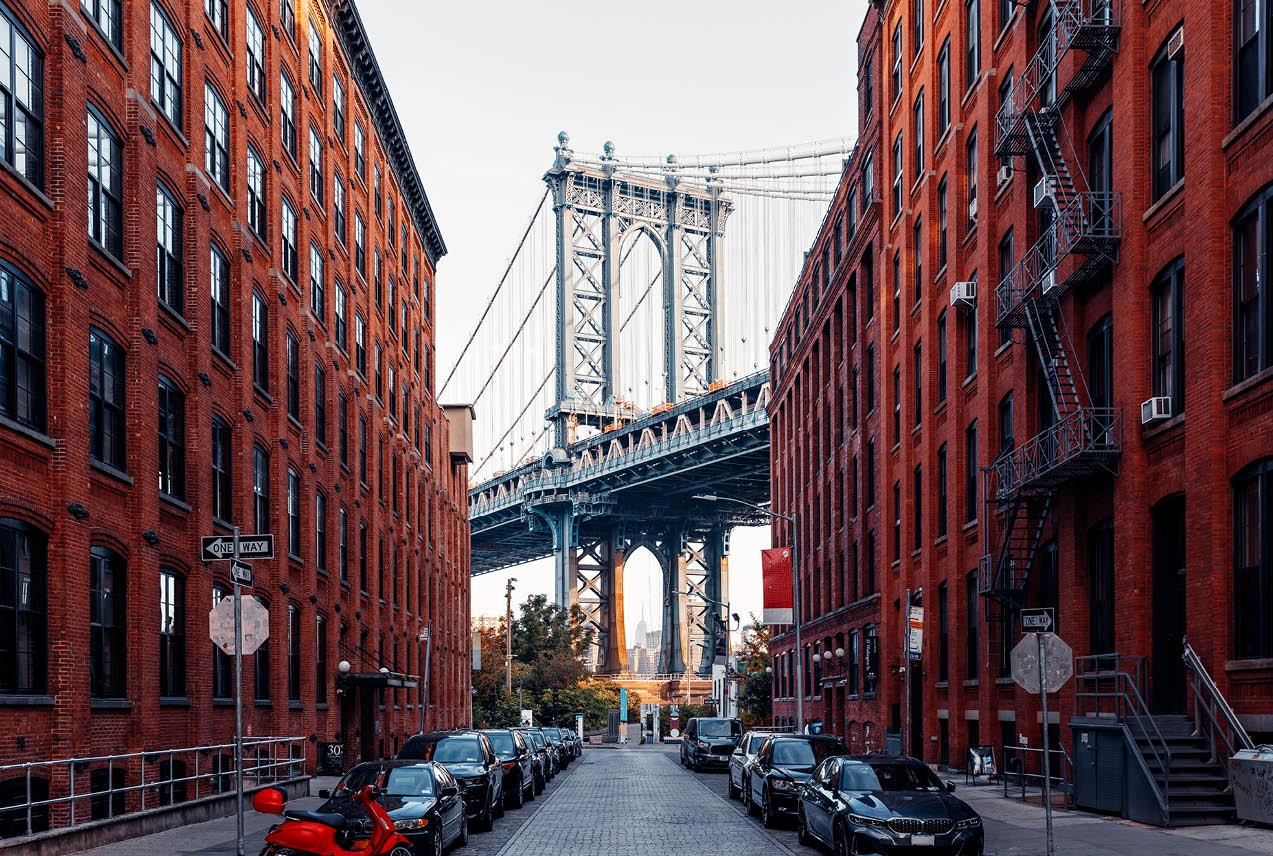 A New York City street lined with parked cars leading up to a bridge in the background