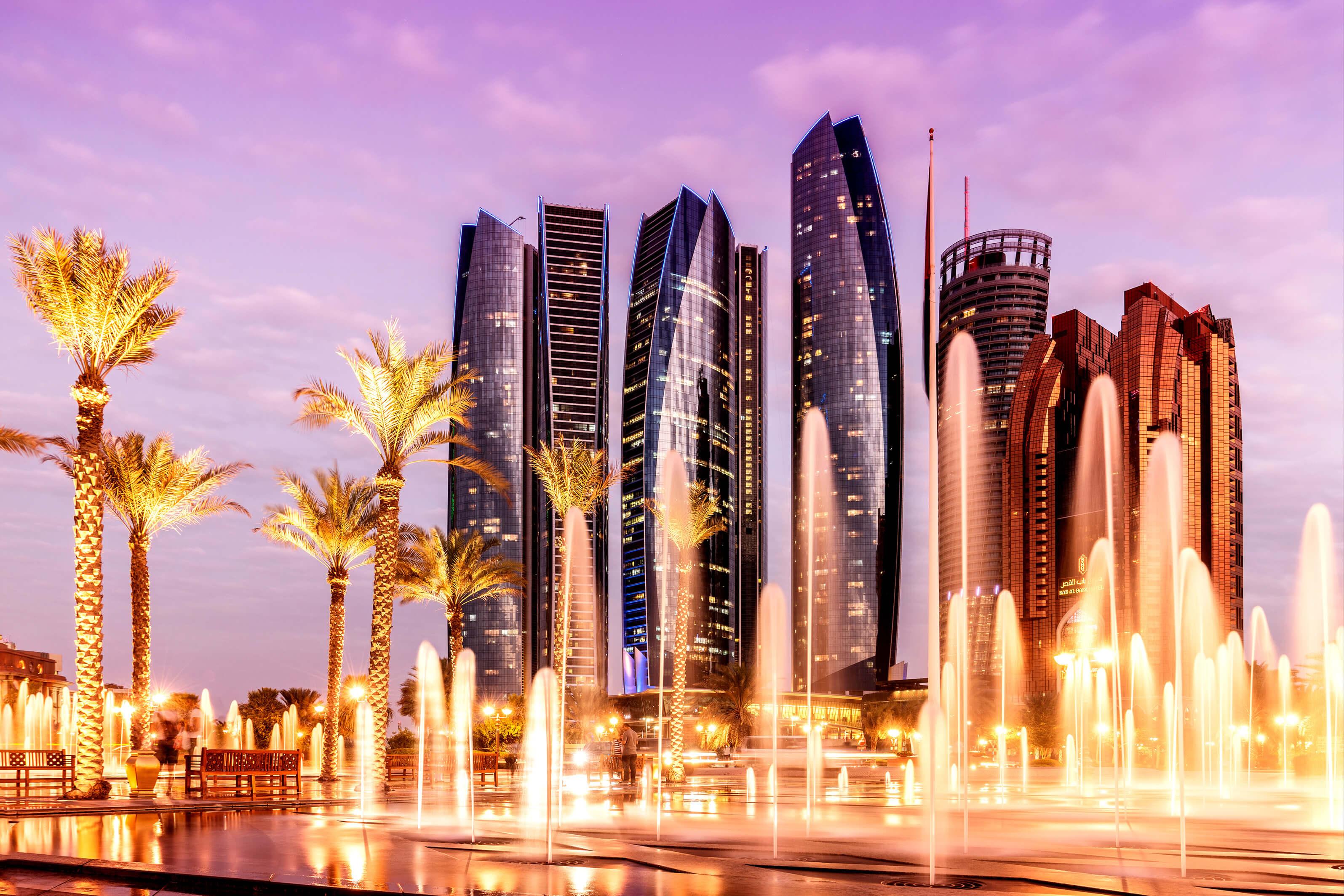 Abu Dhabi skyline at dusk, glittering fountains and trees in foreground 