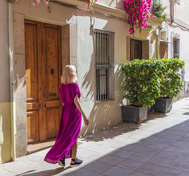 Woman in a fuchsia dress walking down a sunny street with flowering balconies.