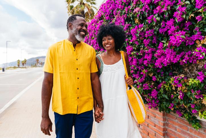 Smiling couple holding hands, walking past a vibrant purple flowering bush.