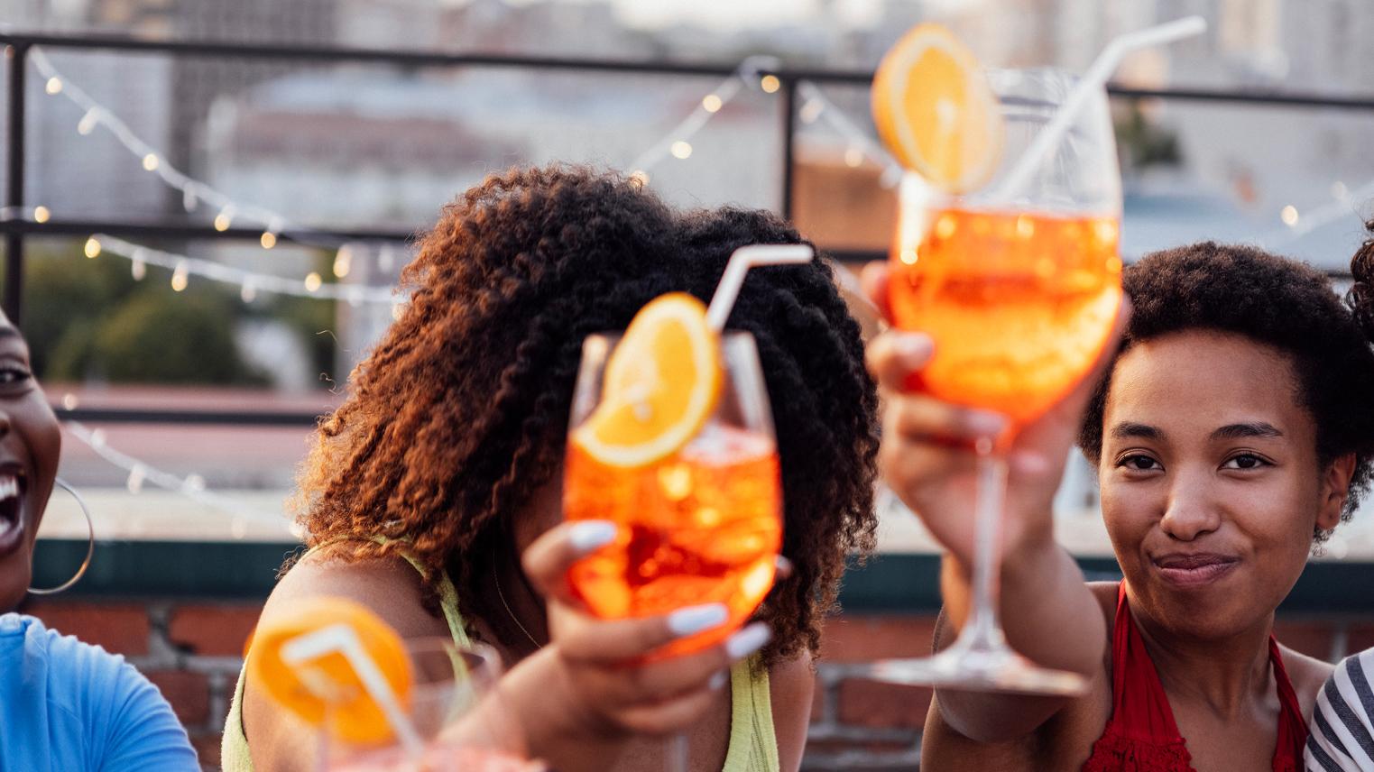 Two women hold up tropical drinks with straws and smile