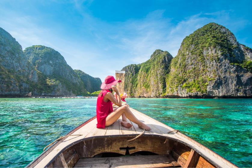 Woman in red dress and hat sitting on boat in clear blue water with cliffs