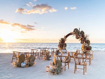 Sunset view of a beach wedding ceremony with wooden chairs, a floral arch, and unique mirrored disco ball decorations.