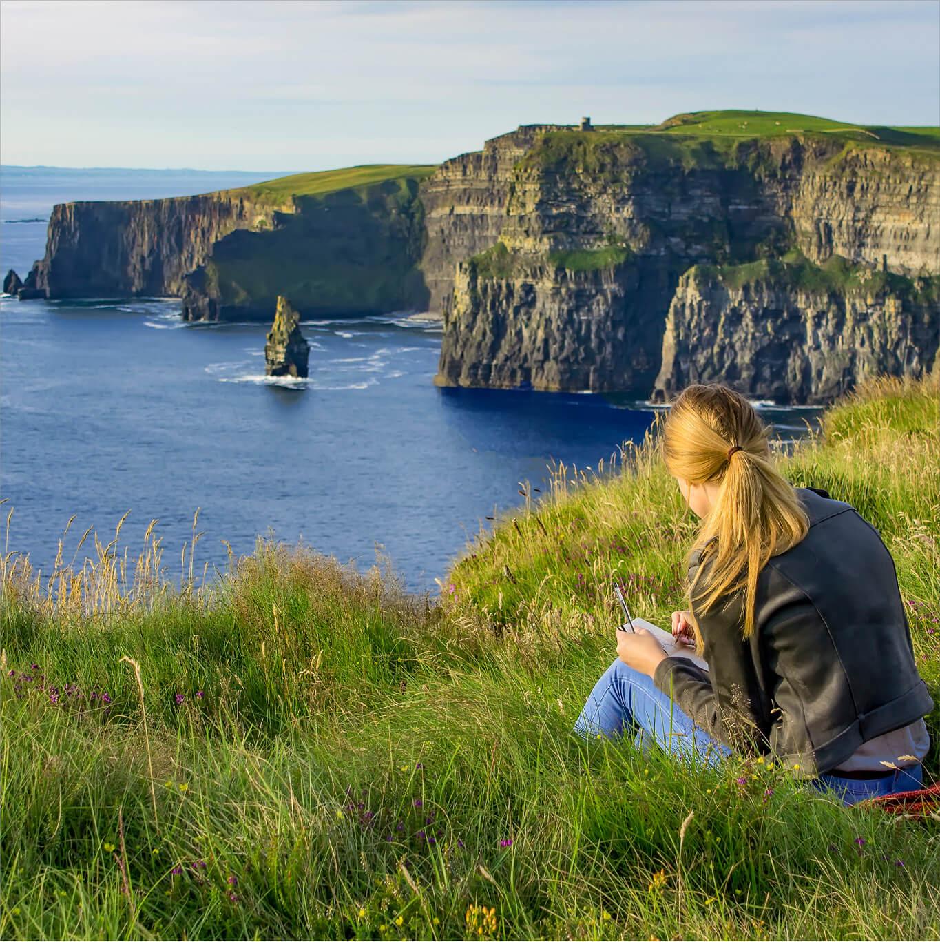 A woman sketches while overlooking the Cliffs of Moher