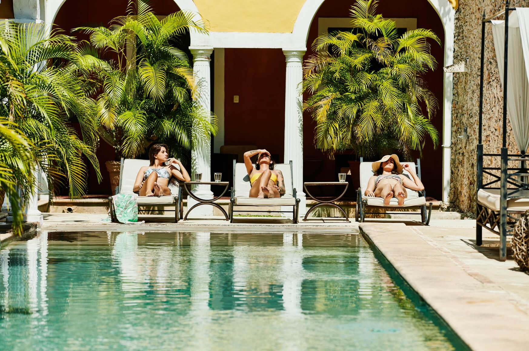 Three women lounge by a pool in a tropical setting