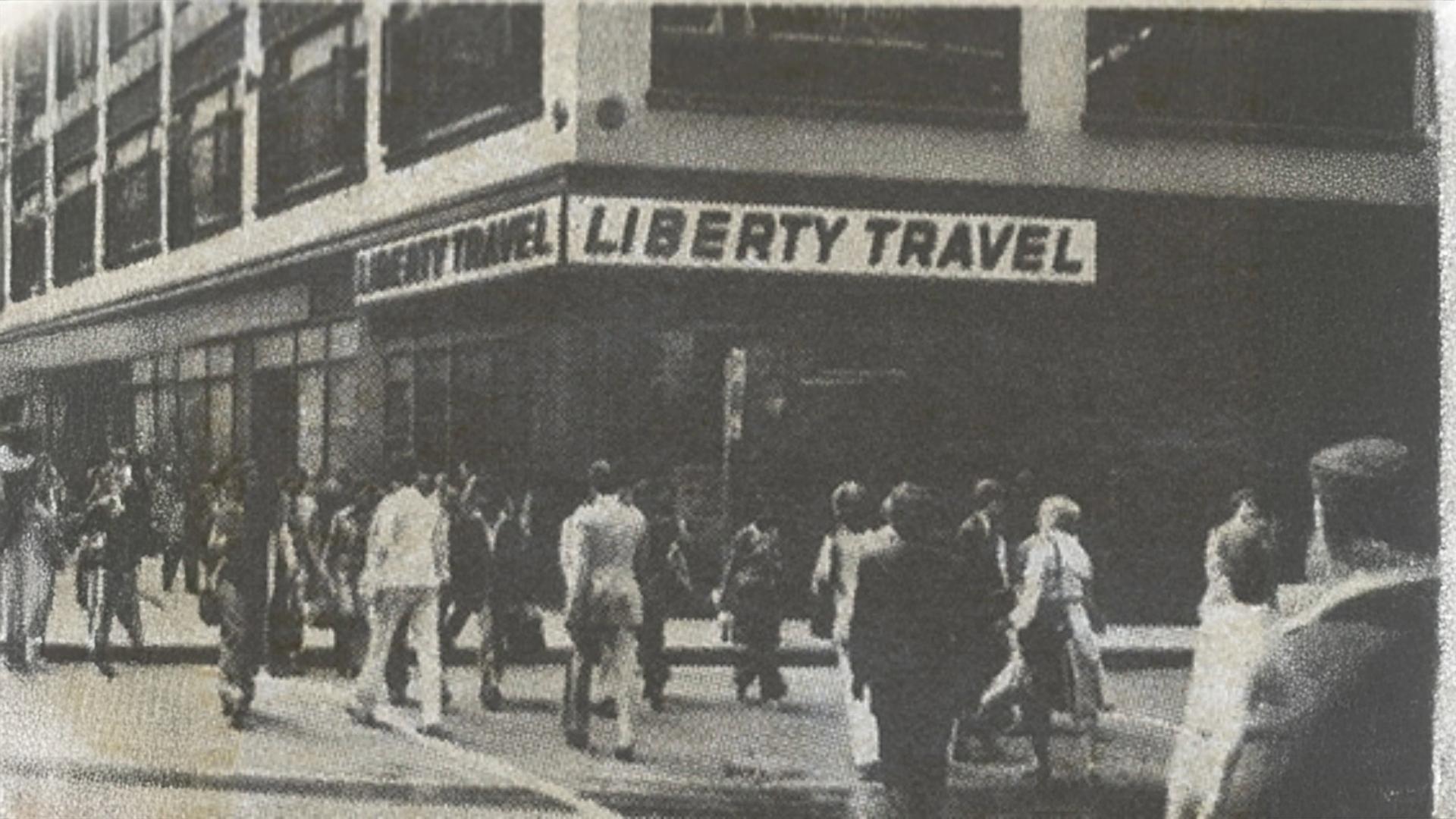 A vintage black and white photograph showing the exterior of a 'Liberty Travel' storefront on a city street corner, with many pedestrians walking in front of the building.