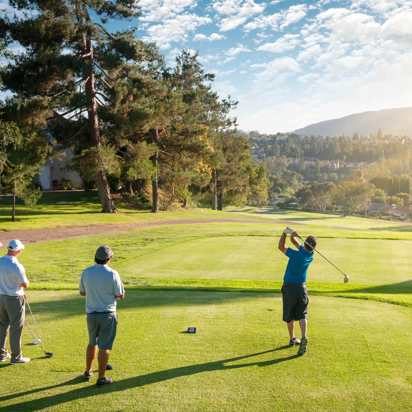 A man takes a golf swing while 2 friends look on