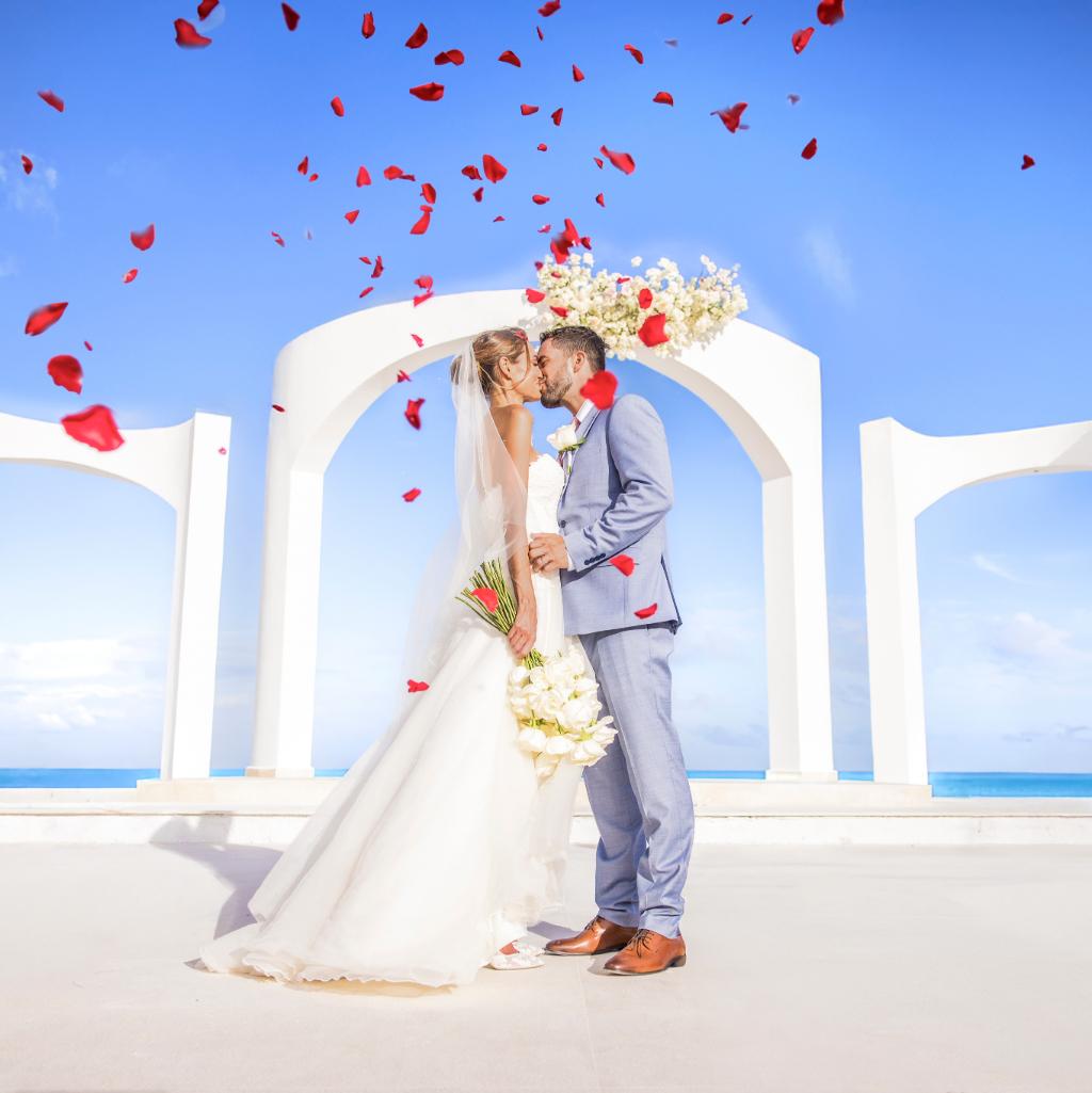 Bride and groom kissing beneath a white ceremony arch overlooking the ocean, with rose petals in the air.