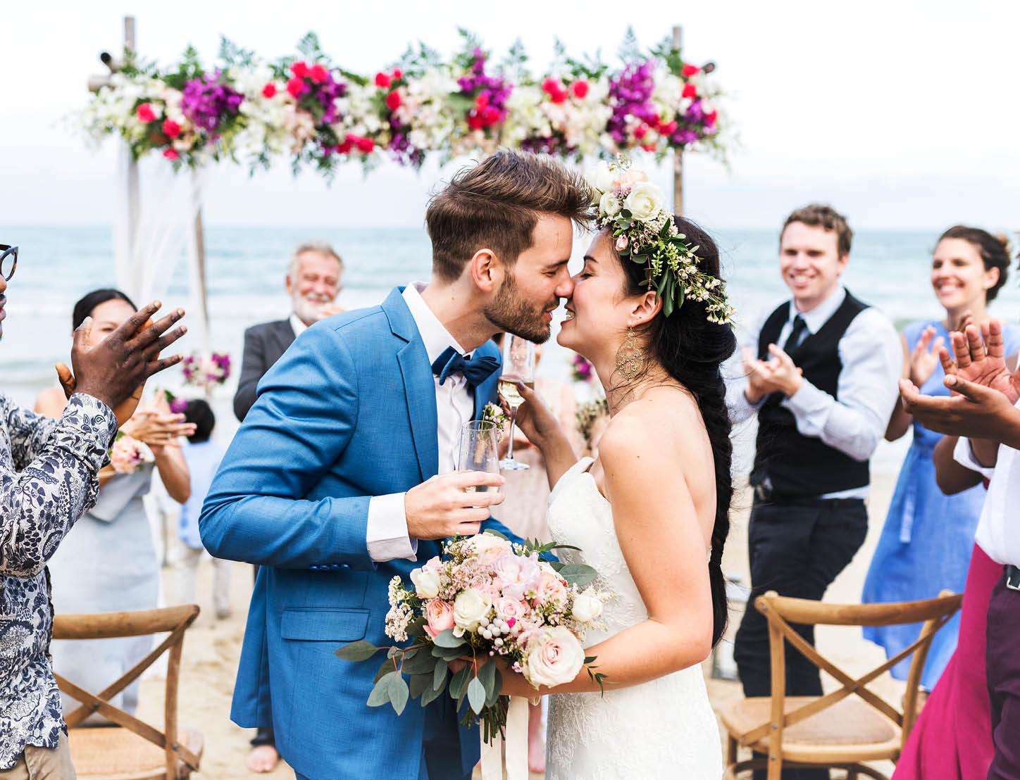 A newlywed couple go in for a kiss with their destination wedding guests in the background
