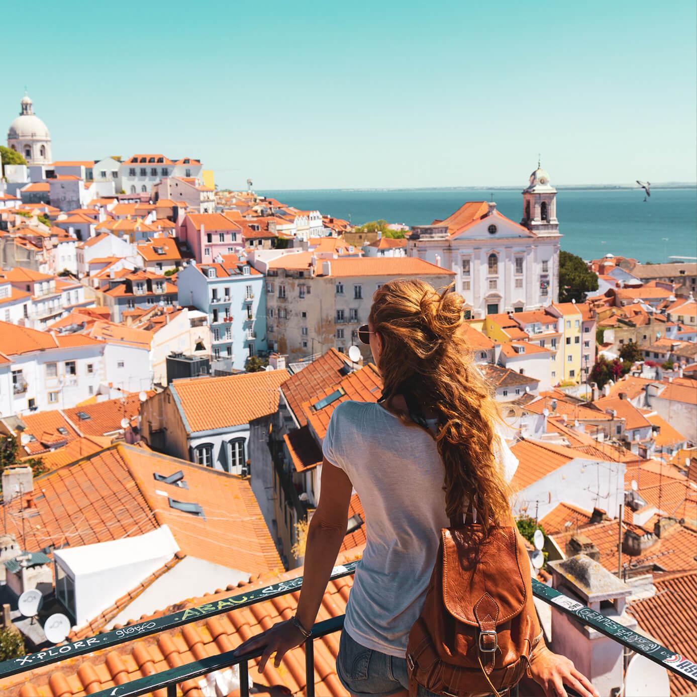 A woman overlooking a scenic view of Lisbon, Portugal, featuring red-tiled rooftops, historic buildings, and the Tagus River