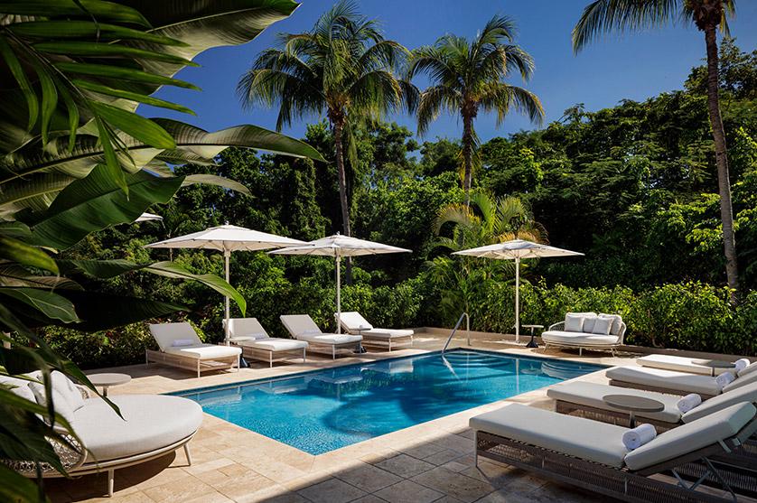 Tranquil resort pool scene featuring a clear blue pool, shaded lounge chairs with white cushions, and lush tropical greenery including palm trees.