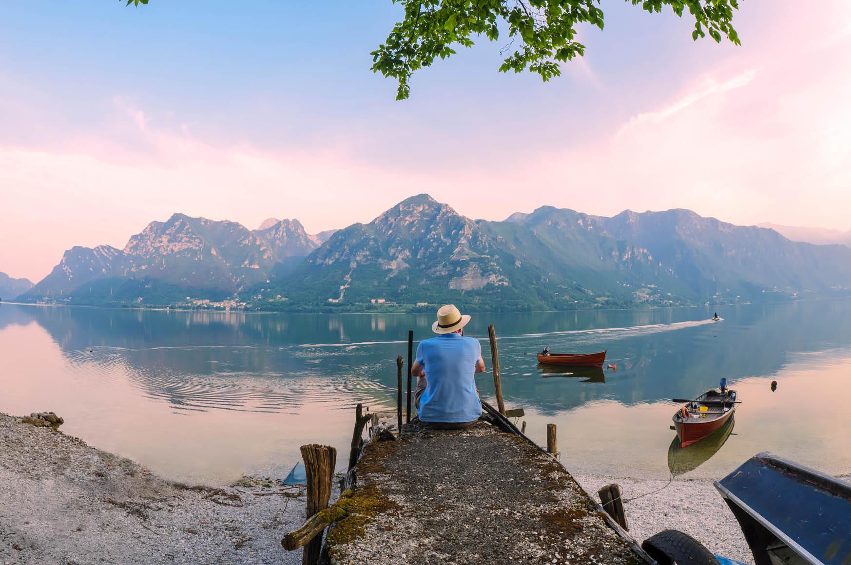 A person sits at the end of a dock, overlooking a lake with mountains in the background
