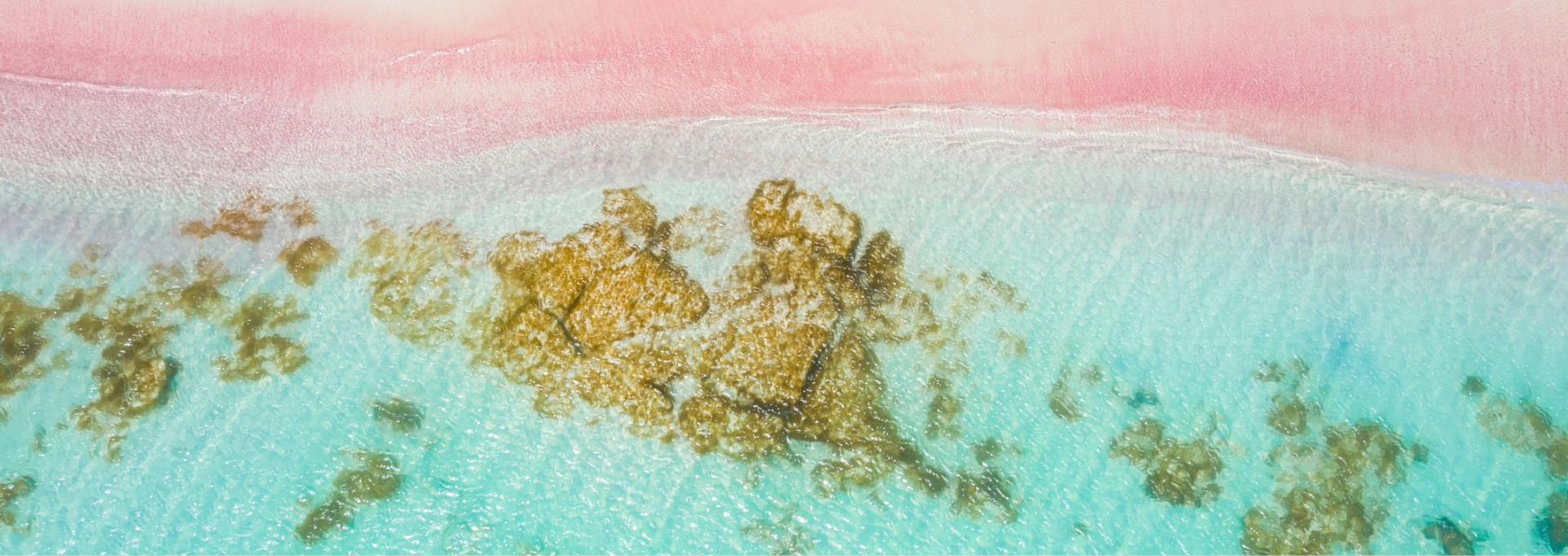 Top-down view of a bright white and pink sand beach next to crystal clear turquoise ocean water, showing the texture of rocks and shallow waves.