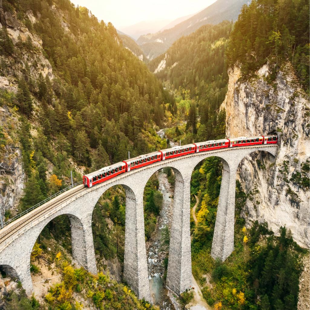 Red scenic train crossing a stone bridge through the Swiss Alps.